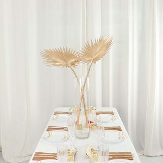 Elegant table setting with gold palm leaf decor and soft white curtains.