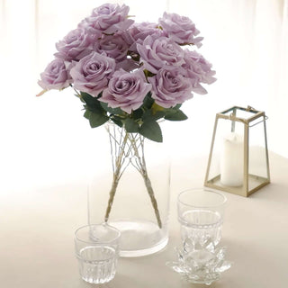 Purple artificial roses in a clear vase with glasses and a candle holder.