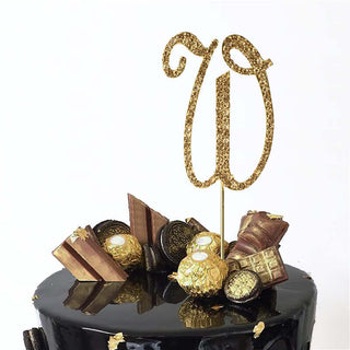 Gold glitter "W" cake topper on a black cake with decorative chocolates.