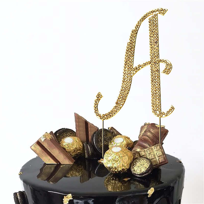 Gold glitter "A" cake topper with decorative elements on a black cake.