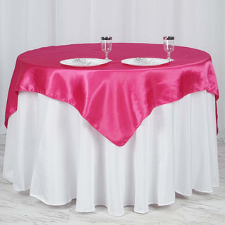 Pink satin tablecloth draped over a white round table with dinnerware and glasses.