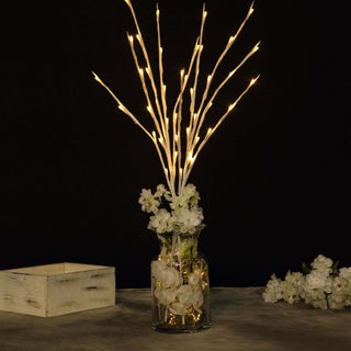 Decorative vase with fairy light branches and white flowers in a dark setting.