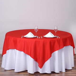 Red tablecloth draped over a round table set for two with glassware and napkins.