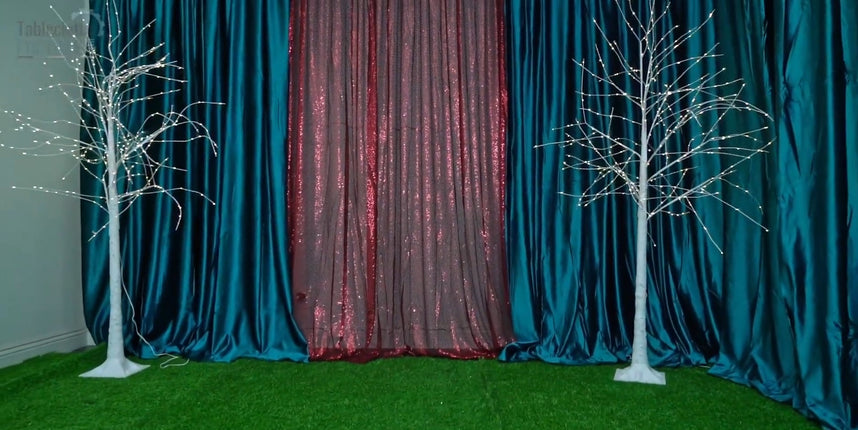 Dual lighted birch trees with teal velvet and burgundy sequin backdrops on green grass carpet.