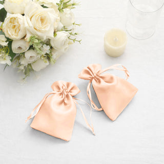 Two peach satin drawstring bags on a table with flowers and a candle.