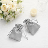Two elegant grey satin drawstring bags on a table with flowers and a candle.