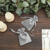 Two silver satin drawstring bags on a wooden table with greenery and candles.