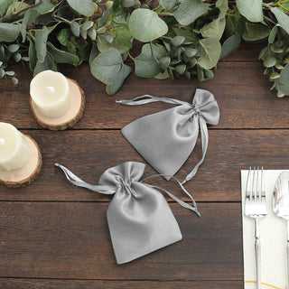 Two silver satin drawstring bags on a wooden table with greenery and candles.