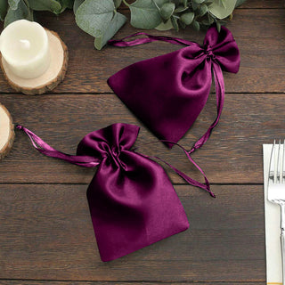 Purple satin drawstring bags on a wooden table with greenery and candles.