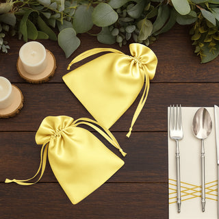 Yellow satin drawstring bags on a wooden table with greenery and tableware.