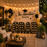 Graduation party backdrop with balloons, lights, and “The Adventure Begins” sign