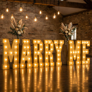 Illuminated “Marry Me” wedding proposal sign with floral arrangements and warm lights