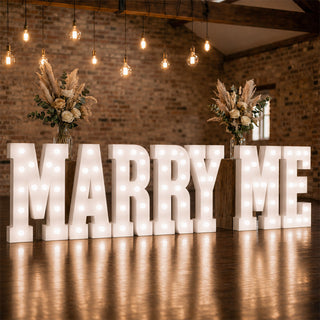 Marquee “MARRY ME” proposal sign with floral arrangements in rustic venue