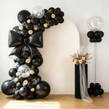 Black and gold balloon arch with roses and a floral arrangement in a studio setting.