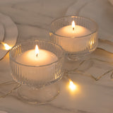 Two lit candles in clear glass holders with warm fairy lights on marble surface