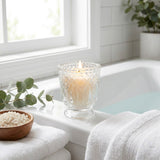 Lit candle in ornate glass holder beside bath towels in a bright bathroom