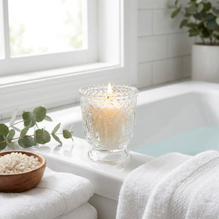 Lit candle in ornate glass holder beside bath towels in a bright bathroom