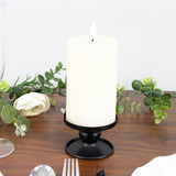 White pillar candle on a black holder with greenery accents on a wooden table.