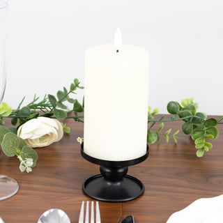 White pillar candle on a black holder with greenery accents on a wooden table.