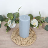 Gray pillar candle on a woven mat surrounded by greenery and flowers.