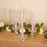 Three lavender candles in clear glass holders on a wooden table with flowers.