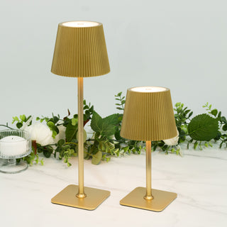 Gold pleated lamps in two sizes on a marble surface with greenery backdrop.