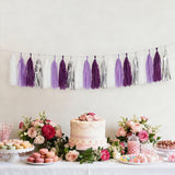 Purple and silver tassel garland above a decorated dessert table with cake and flowers.