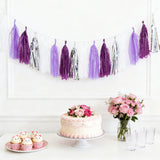 Purple and white tassel garland with cake, cupcakes, and flowers on a table.