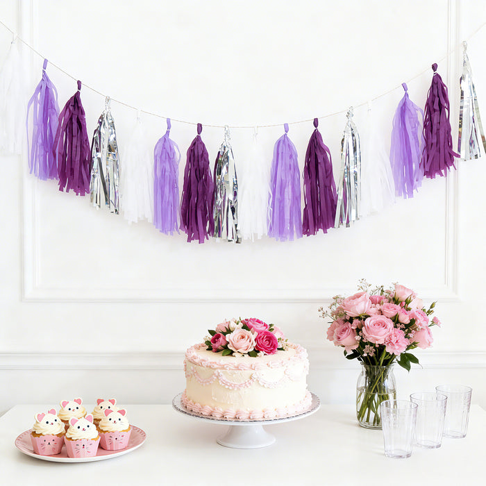 Purple and white tassel garland with cake, cupcakes, and flowers on a table.