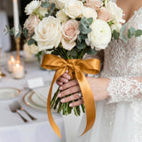 Bride holding white and blush rose bouquet with gold ribbon at wedding table