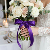 Bride holding white and blush rose bouquet with purple ribbon and lace dress