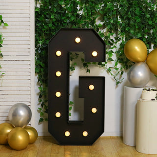 Decorative light-up letter "G" surrounded by balloons and greenery.