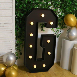Decorative marquee letter "G" with lights, set against greenery and balloons.