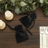 Black satin drawstring bags on a wooden table with greenery and candles.