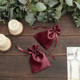 Burgundy satin drawstring bags on a wooden table with greenery and candles.