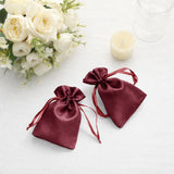 Two burgundy satin drawstring bags on a table with flowers and a candle.