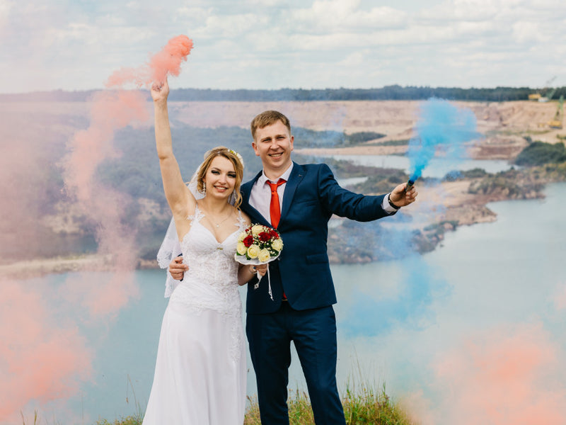 Bride and groom holding colored smoke flares by a lake
