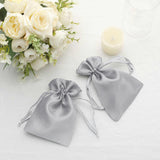 Silver satin drawstring bags on a table with flowers and a candle.