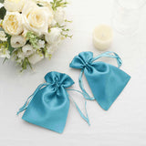 Two elegant teal satin drawstring bags on a soft white surface with flowers and a candle.