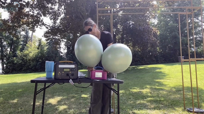 Person using an electric balloon pump to inflate two dusty sage green balloons outdoors.