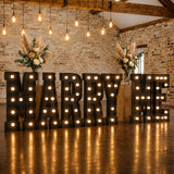 Illuminated “Marry Me” wedding sign with floral arrangements in rustic venue