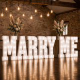 Marquee “MARRY ME” proposal sign with floral arrangements in rustic venue