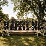 Outdoor wedding marquee sign spelling “Marry Me” with floral decor and candles