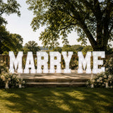 Elegant outdoor wedding sign reading “MARRY ME” with flowers and lights