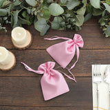 Two pink satin drawstring bags on a wooden table with greenery and candles.