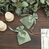 Green satin drawstring gift bags on a wooden table with candles and greenery.