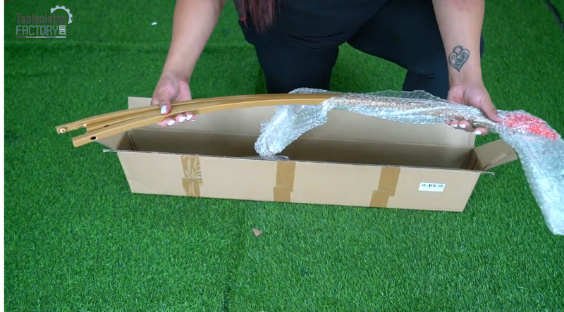 Person unwrapping gold arched frame pieces from a cardboard box, preparing to assemble a Chiara backdrop stand.
