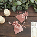 Rose satin drawstring gift bags on a wooden table with greenery and candles.