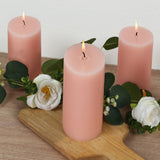 Three pink candles surrounded by greenery and flowers on a wooden surface.