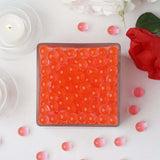 Orange gel beads in a square container surrounded by flowers and candles.